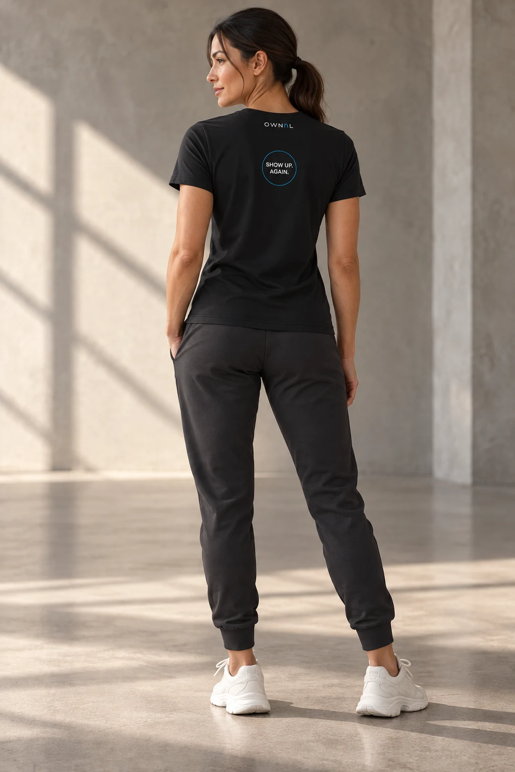 Female model showing a small SHOW UP AGAIN back print on a black OWNAL tee